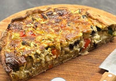Eine halbe Quiche mit gebräunter Decke. Viel kleingeschnittenes Gemüse ist an der Schnittseite zu sehen. Die Quiche liegt auf einem Holzbrett.