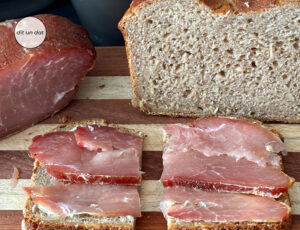 Im Vordergrund eine Scheibe Brot mit Schinken. Im Hintergrund links eine Teilstück des Schinkens und rechts im Hintergrund ein aufgeschnittenes Kastenbrot.