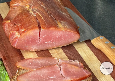 Ein länglicher Rohschinken auf einem Holzbrett. Rechts danemben ein Fleischmasser, im Vordergrund eine Scheibe Brot, belegt mit Schinken